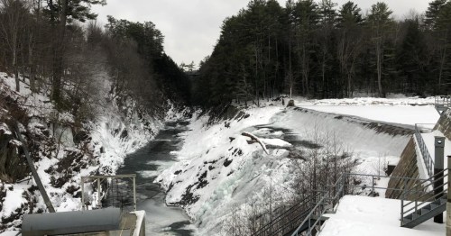 A snowy landscape featuring a partially frozen river and a dam, surrounded by trees in a winter setting.