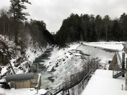 A snowy river flows through a rocky landscape, with a dam and trees lining the banks under a cloudy sky.