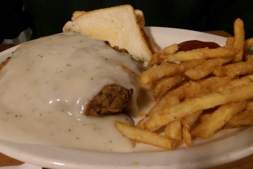 A plate featuring a breaded meat patty smothered in white gravy, served with fries and two slices of bread.