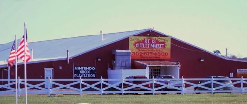 Red barn-style building with a sign for PT 13 Outlet Market, featuring gaming consoles and American flags in the foreground.