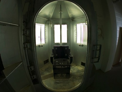 A dimly lit execution chamber with a central chair, surrounded by windows and industrial features.
