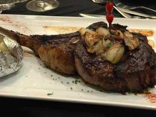 A large, grilled tomahawk steak topped with sautéed onions and garnished with a cherry tomato on a white plate.