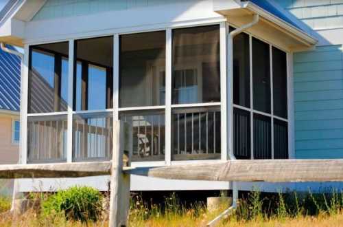 A screened porch attached to a light-colored house, surrounded by grass and a wooden fence.