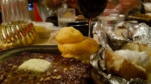 A plate with a juicy steak topped with butter, a baked potato in foil, and a fluffy roll, with drinks in the background.