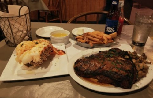 A plate with grilled steak, lobster tail, mushrooms, and fries, alongside a drink and dipping sauce on a restaurant table.
