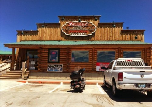 A rustic log cabin-style food mart with a green roof, featuring a motorcycle and a pickup truck parked outside.