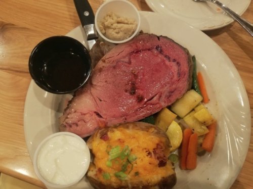 A plate featuring prime rib, baked potato with toppings, mixed vegetables, and dipping sauces.