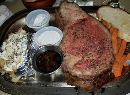 A plate featuring a large prime rib steak, baked potato, carrots, dipping sauces, and a slice of bread.