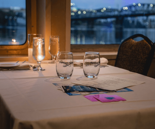 A table set for dining with two glasses, a candle, and a view of a river and bridge in the background.
