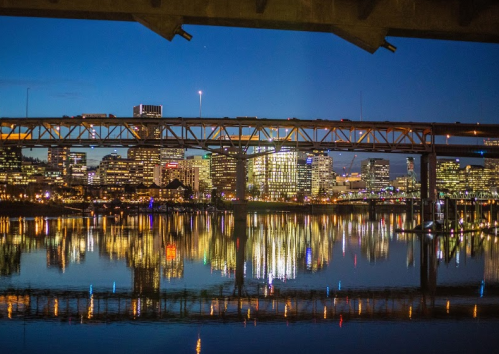 City skyline at night with illuminated buildings reflecting on the water beneath a bridge.