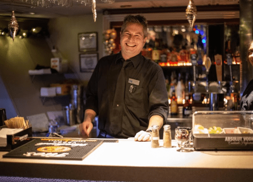 A smiling bartender stands behind a bar, with drinks and garnishes visible in a lively, dimly lit setting.