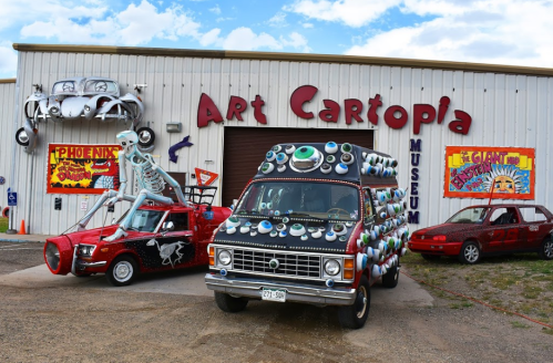 Colorful, artistic vehicles outside the Art Cartopia Museum, featuring a skeleton car and an eye-covered van.
