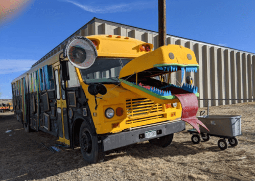 A colorful bus with a large, playful dragon face and a long tongue, parked in a rural area.