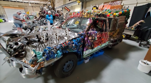 A creatively decorated car covered in colorful toys, metal parts, and sculptures, displayed in a large indoor space.