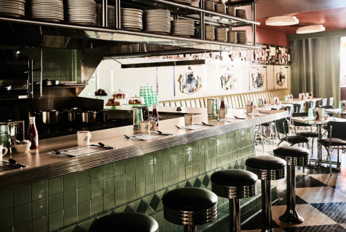 A retro diner interior featuring a green tiled counter, chrome stools, and colorful artwork on the walls.