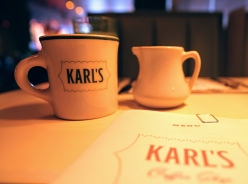 A coffee cup and a cream pitcher on a table, with a menu featuring "Karl's" in the background.