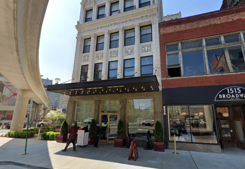 Historic building entrance with awning, potted plants, and nearby urban architecture. Bright, sunny day.