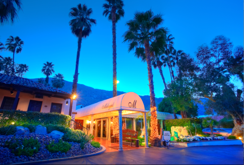 A charming entrance to a hotel surrounded by palm trees, illuminated at dusk with a mountain backdrop.