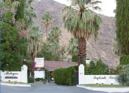 Entrance to a restaurant and inn surrounded by palm trees and mountains. Signage visible at the entrance.
