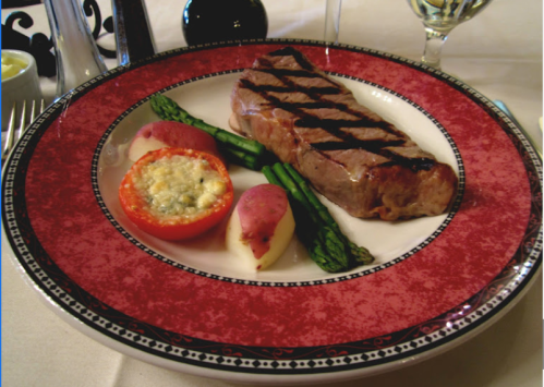 A plated meal featuring grilled steak, asparagus, a stuffed tomato, and red potatoes on a decorative red plate.