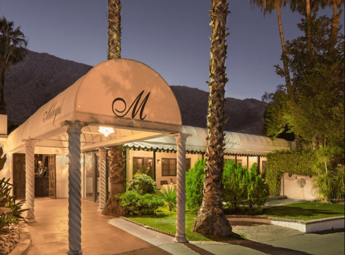A stylish entrance to a restaurant with palm trees, a canopy, and mountains in the background, illuminated at dusk.