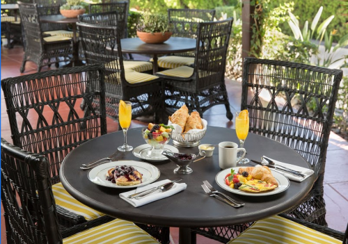 A beautifully set outdoor table with breakfast dishes, fruit, croissants, and drinks in a sunny garden setting.