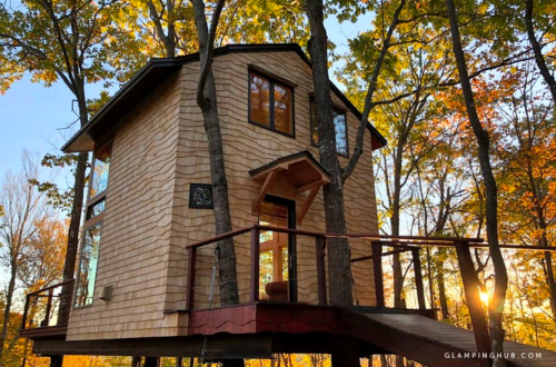 A cozy treehouse with large windows, nestled among autumn trees, featuring a wooden deck and a warm sunset glow.