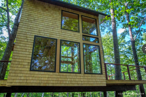 A modern wooden treehouse with large windows, surrounded by lush green trees and a clear blue sky.