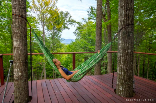 A person relaxes in a green hammock between two trees on a wooden deck, surrounded by lush greenery and mountains.