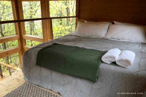 Cozy bedroom with a large bed, green blanket, and towels, surrounded by large windows and forest views.