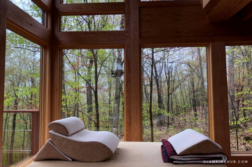 Cozy interior with wooden beams, a bed, and large windows overlooking a lush forest.