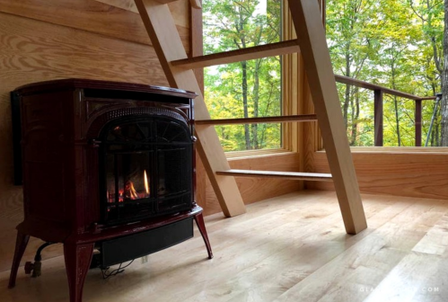 Cozy interior with a red stove, wooden stairs, and large windows showcasing a forest view.
