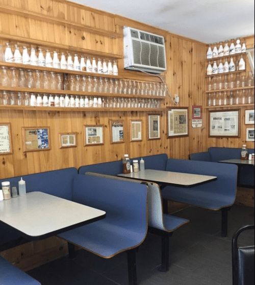 Cozy diner interior with wooden walls, blue booths, and shelves lined with bottles and framed newspaper clippings.