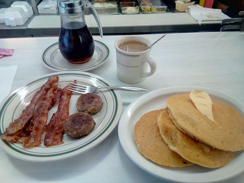 A plate with pancakes, bacon, and sausage, alongside a coffee cup and a carafe of syrup on a diner table.