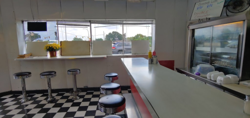 A diner interior with a checkered floor, bar stools, a counter, and a window view. Flowers and condiments are on the counter.