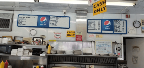 Interior of a diner with menus on the wall, cash-only signs, and a grill area with condiments visible.
