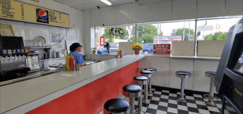 A diner interior with a counter, stools, and a customer. Brightly lit with a view of the street outside.