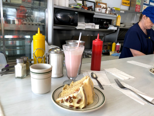 A slice of cake on a plate with a cup of coffee and a milkshake on a diner table, with condiments in the background.