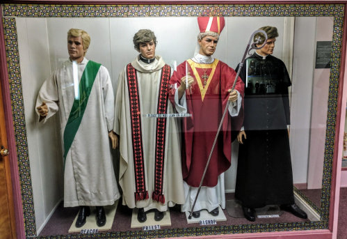 Four mannequins dressed in religious attire: a deacon, priest, bishop, and a figure in a black robe.
