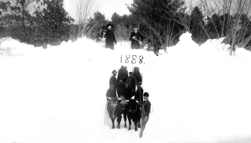A historic black-and-white photo of people and horses in deep snow, dated 1888, with trees in the background.