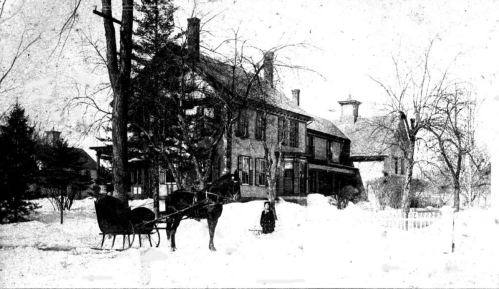 A vintage winter scene featuring a horse-drawn sleigh in front of a large house surrounded by snow-covered trees.