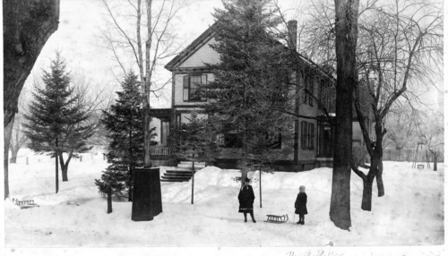 A vintage black-and-white photo of a snowy scene with a large house and two children near a sled.