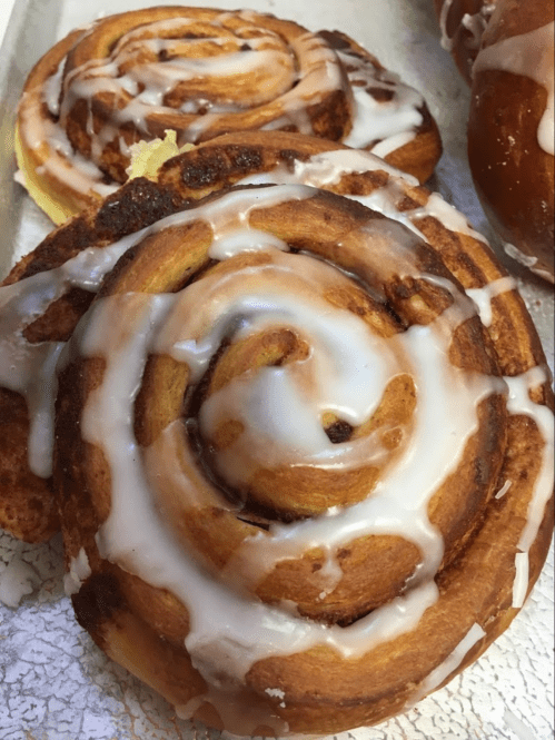 Close-up of a cinnamon roll drizzled with icing, resting on a tray with another roll in the background.