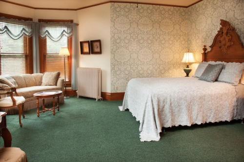 Cozy bedroom with a large bed, vintage furniture, and patterned wallpaper, featuring soft lighting and a green carpet.