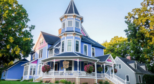 A colorful Victorian-style house with a turret, surrounded by trees and flowers, featuring a welcoming porch.