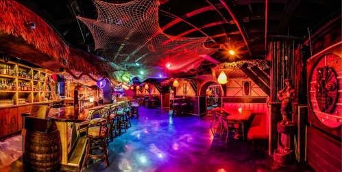 Vibrant bar interior with colorful lights, tropical decor, and a lively atmosphere, featuring a thatched roof and netting.