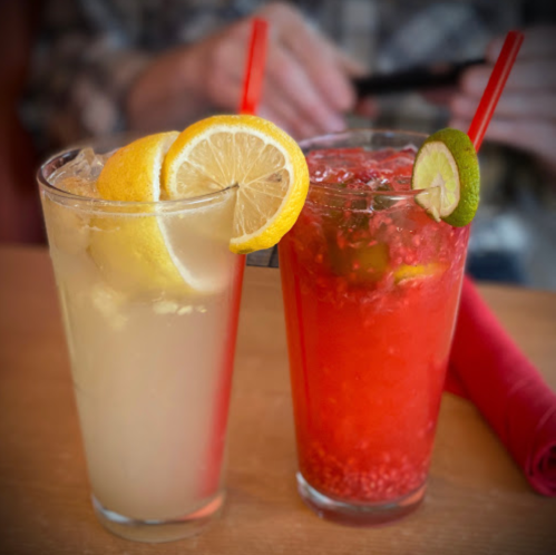 Two colorful cocktails on a table: one with lemon slices and the other with lime and raspberries, both with straws.