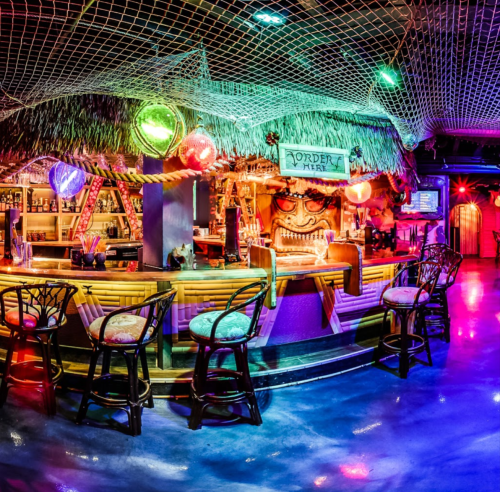 A vibrant tiki bar with colorful lights, thatched decor, and a lively atmosphere, featuring a unique tiki mask centerpiece.