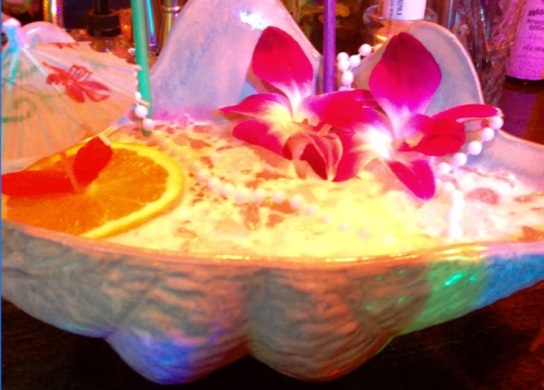A colorful cocktail in a shell-shaped glass, garnished with orange slices and pink flowers.