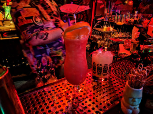A vibrant cocktail with an orange slice sits on a bar, surrounded by colorful decor and another drink in the background.